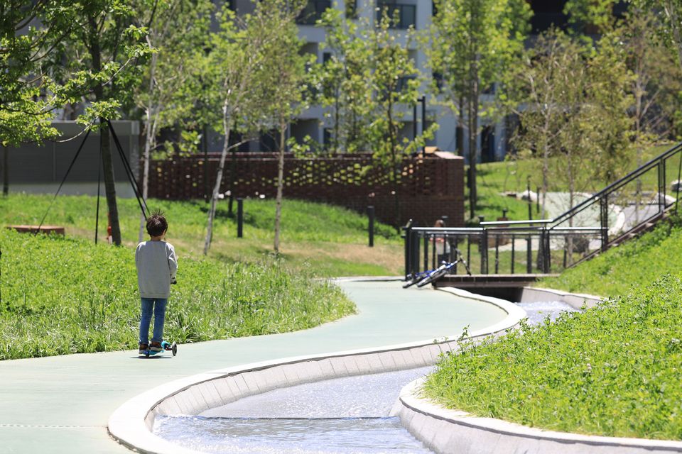 Außenaufnahme eines begrünten Parkwegs mit geschwungener Wasserführung, offenen Aufenthaltsflächen und angrenzenden Wohngebäuden im Hintergrund.