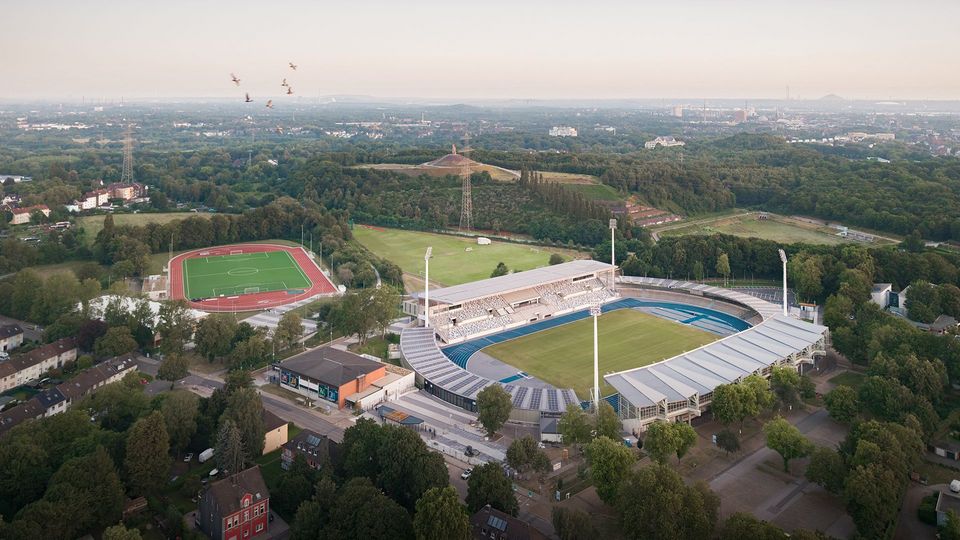 Innenaufnahme eines Tribünenbereichs mit farblich abgesetzten Sitzschalen, verglaster Stirnseite und Blick auf die Laufbahn.