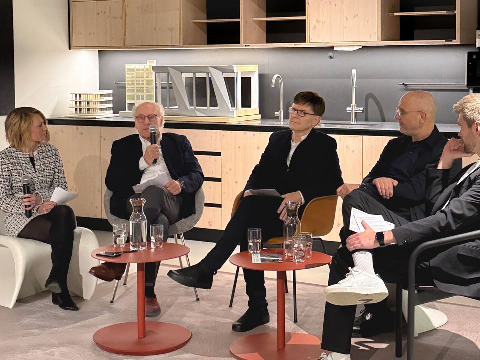 Innenaufnahme einer moderierten Gesprächsrunde mit mehreren Personen, Sitzmöbeln und Modellbauten im Hintergrund in einem modern gestalteten Raum.