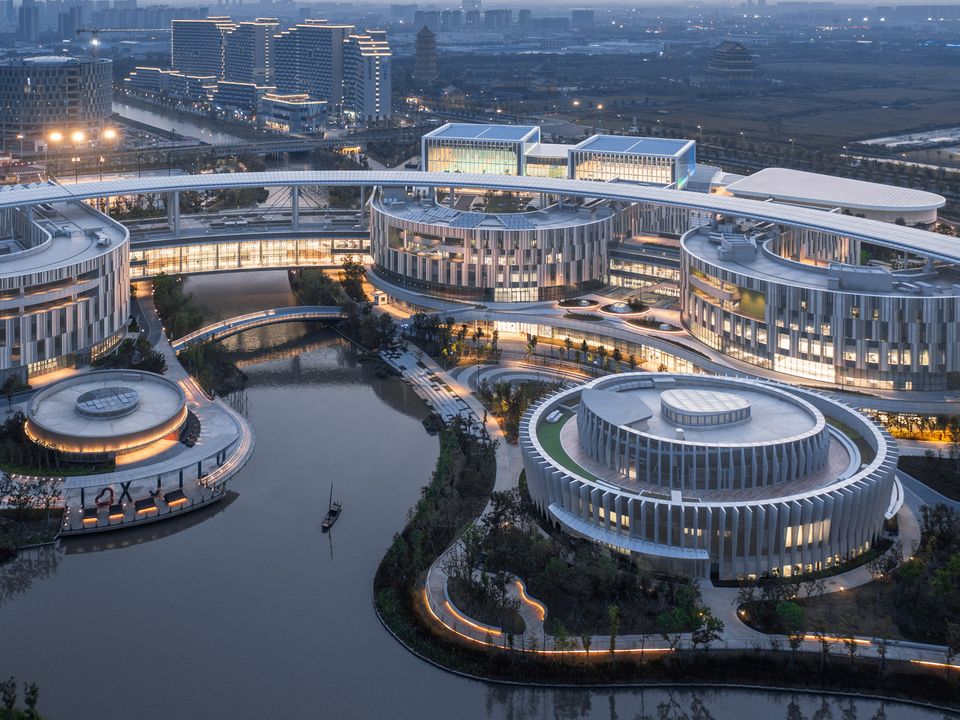 Exterior shot of a modern educational campus with circular and linear buildings, illuminated façades and a central watercourse as a connecting landscape feature.