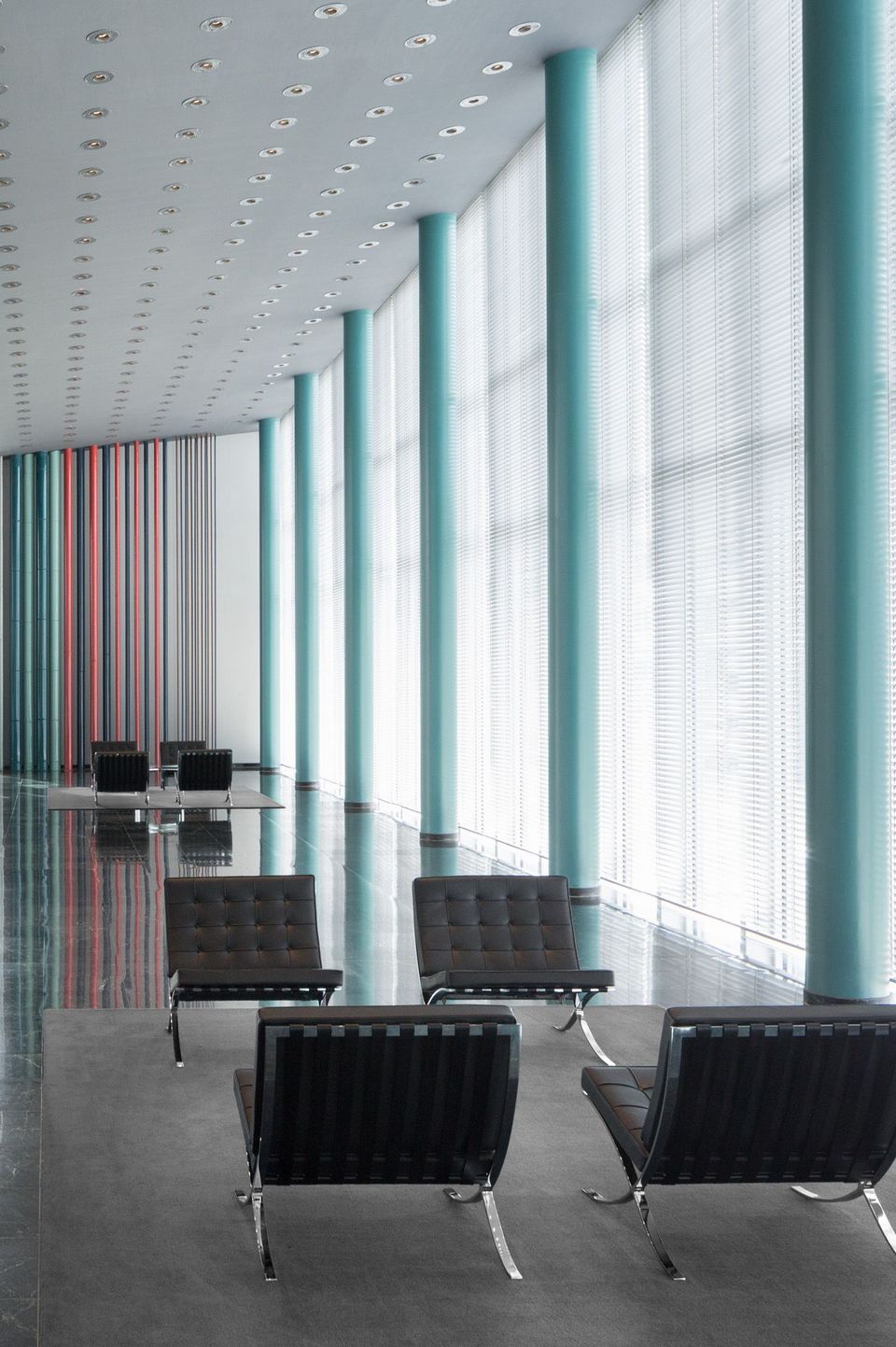 Innenansicht eines großzügigen Foyers mit hohen türkisfarbenen Säulen, bodentiefen Fenstern und schwarzen Ledersesseln im minimalistischen Stil
