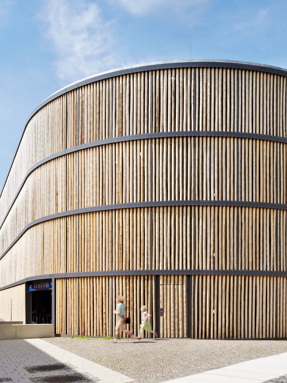 Ein-/Ausfahrt eines Parkhauses mit geschwungener Fassade aus vertikalen Holzlamellen, markantes Design mit natürlicher Materialität