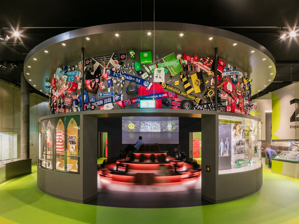Innenaufnahme eines runden Ausstellungsraums mit überhöhtem Präsentationspodest und farbintensiver Wandinstallation aus Fanartikeln, die die emotionale Erlebniswelt des Fußballs visualisiert.