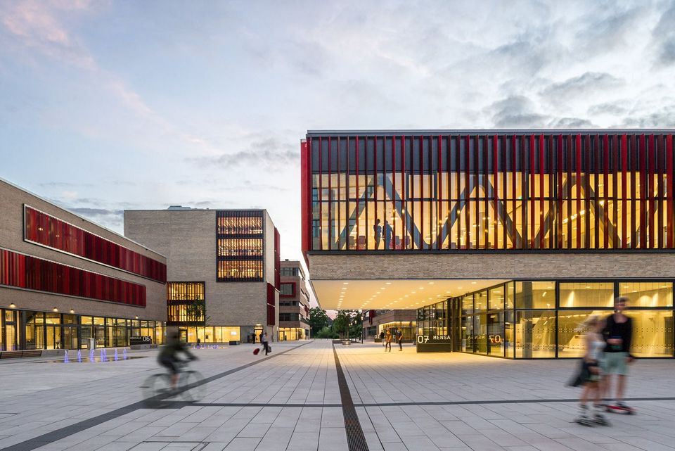 Außenaufnahme eines weitläufigen Campusplatzes, der von verglasten Neubauten mit beleuchteten Innenräumen umgeben ist.