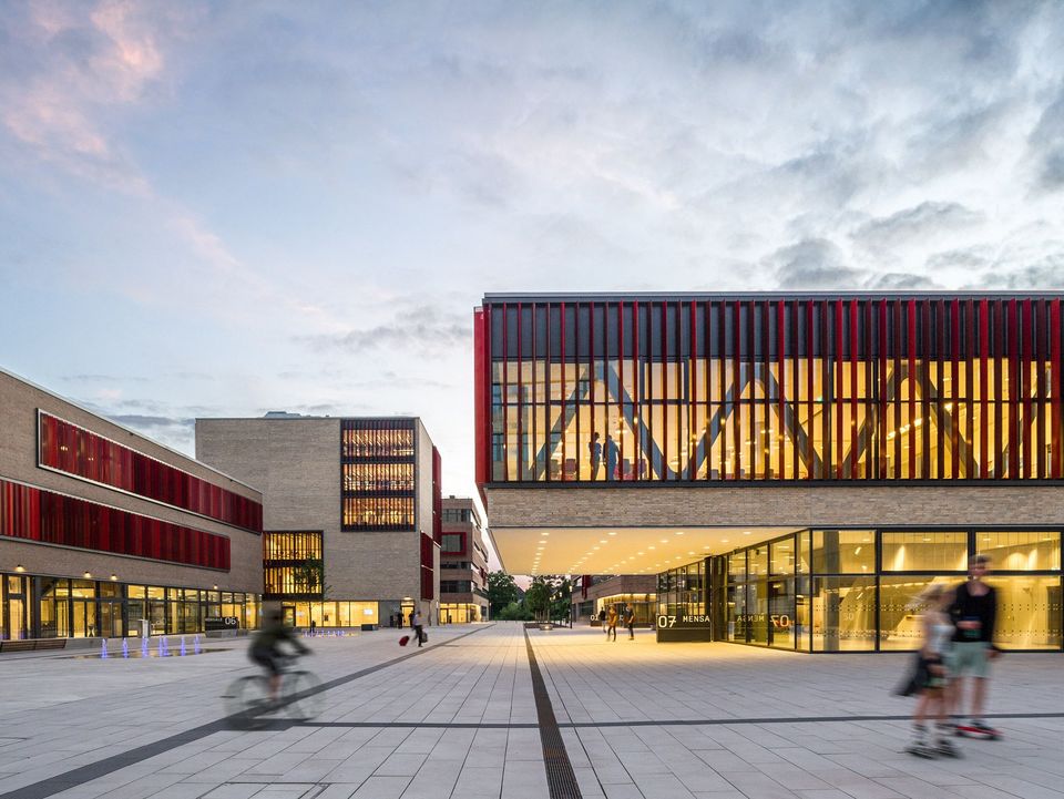 Außenaufnahme eines weitläufigen Campusplatzes, der von verglasten Neubauten mit beleuchteten Innenräumen umgeben ist.