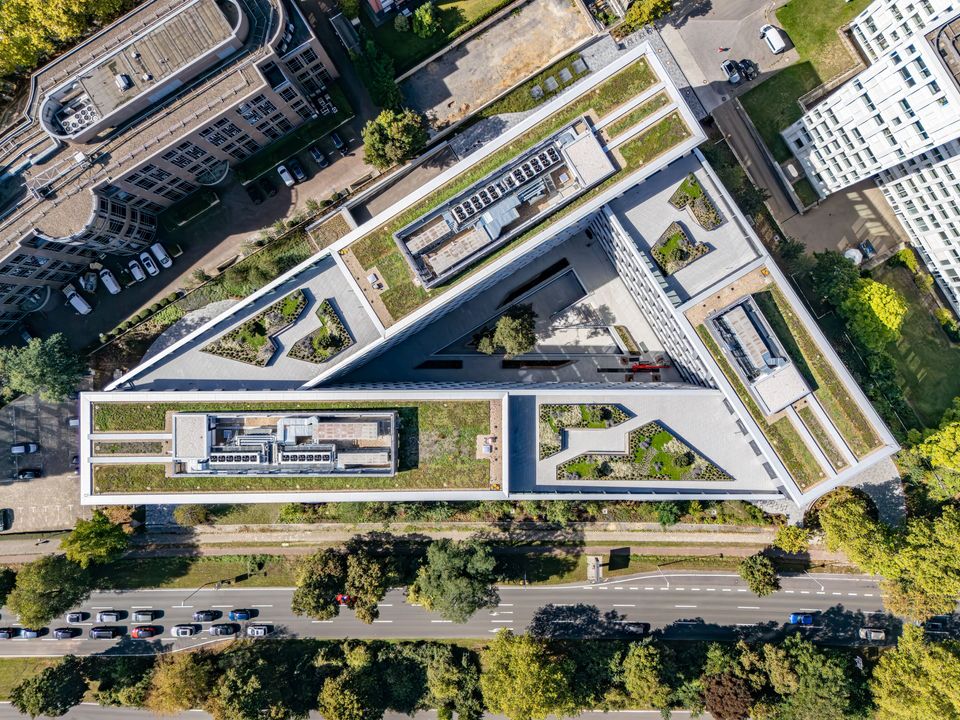 Luftaufnahme eines dreiecksförmigen Bürogebäudes mit weitläufigen Dachgärten, begrünter Fassadenabstufung und umliegender städtischer Infrastruktur.