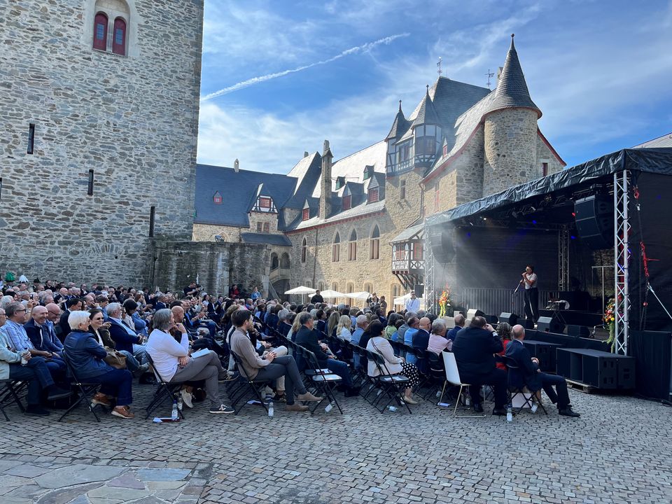 Außenaufnahme einer Publikumsveranstaltung im Burghof mit historischer Architektur, großer Zuschauerfläche und Bühnenaufbau an der Seitenwand.