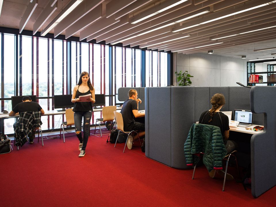 Innenaufnahme einer Bibliothek mit offenen und abgeschirmten Arbeitsplätzen, an denen mehrere Personen konzentriert arbeiten.