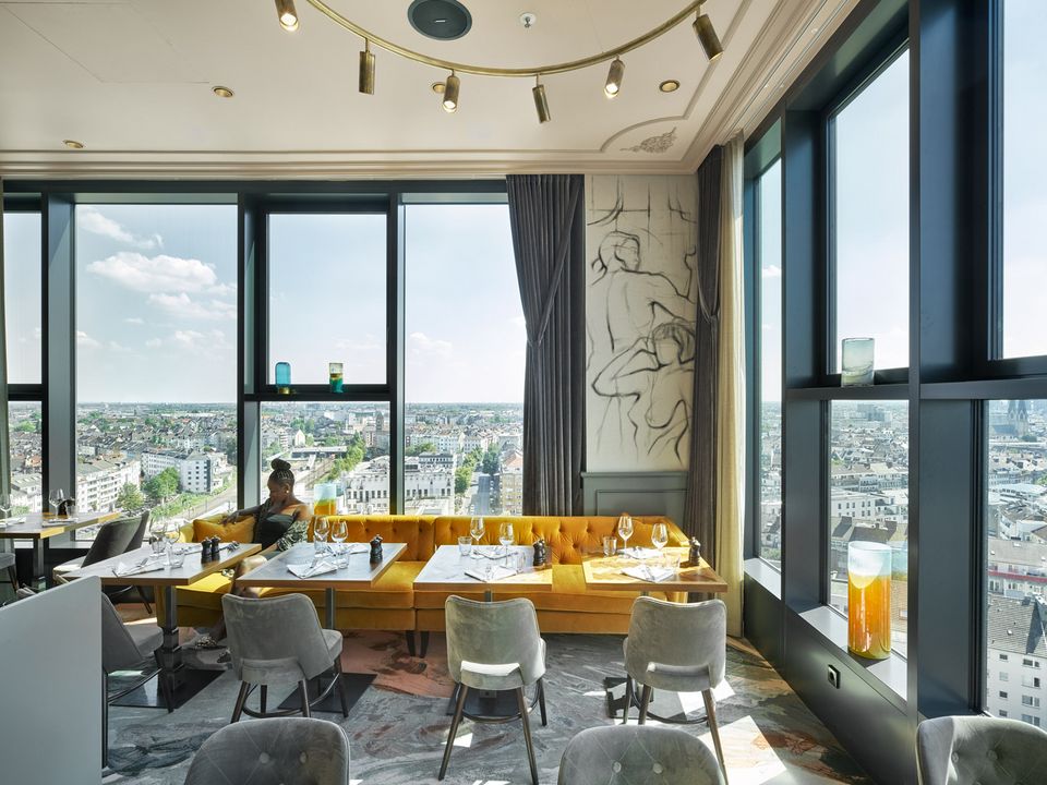 Restaurantbereich mit Panoramafenstern und Blick über die Stadt, gelbe Sitzbank und elegante Tischgestaltung in modernem Interieur