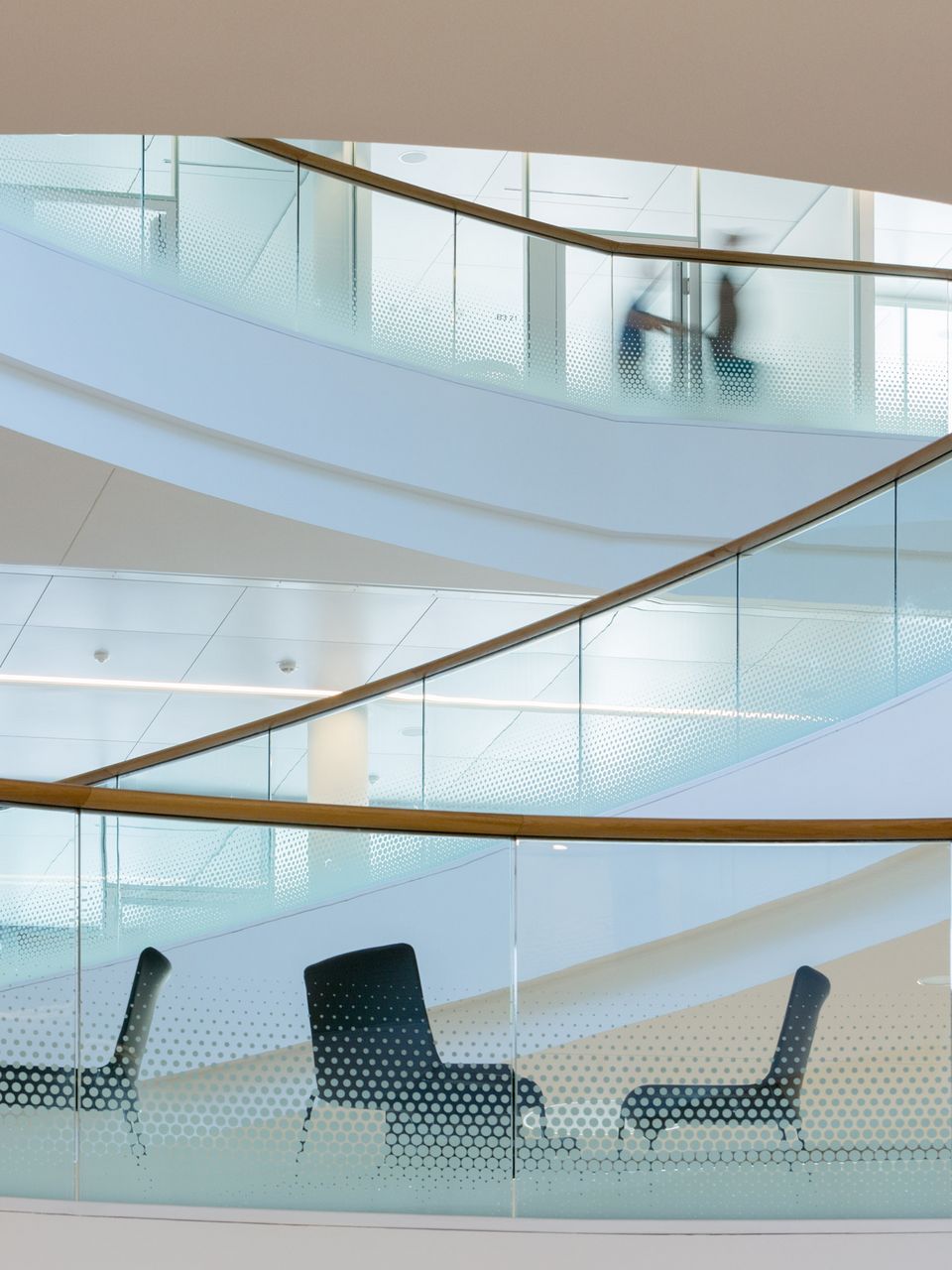 Innenaufnahme eines offenen Galeriegeschosses mit gläsernen Brüstungen und Sitzbereichen entlang der umlaufenden Balustrade.