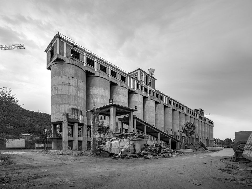 Historische Außenaufnahme eines industriellen Silogebäudes mit offenen Stahlkonstruktionen, Anbauten und unbefestigtem Gelände im Vordergrund.