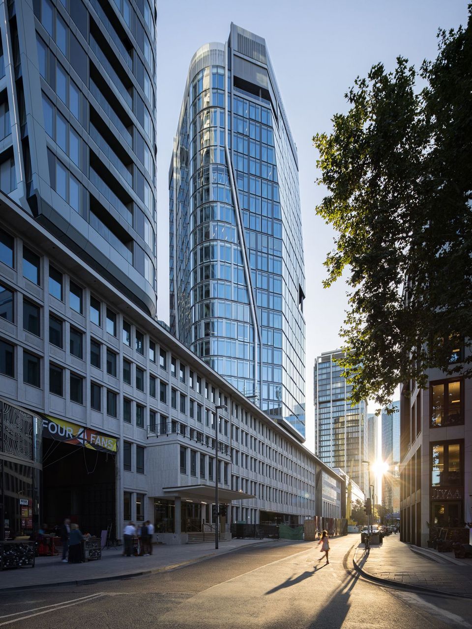 Straßenperspektive im Hochhausquartier mit dominantem Turm, durchgehender Glasfassade, flankierenden Gebäuden und langer Sichtachse in Richtung Sonnenstand.