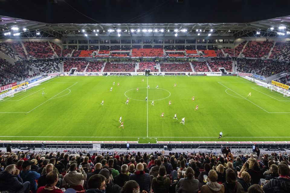 Spielszene im beleuchteten Stadion bei Nacht, volles Haus mit Zuschauern und grünem Spielfeld, klare Sicht auf Tribünen und Dachstruktur