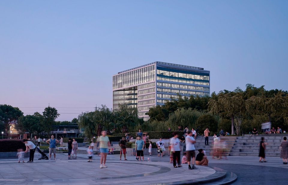 Außenaufnahme des SAIC Volkswagen Technical Centre in Abendstimmung mit leuchtender Glasfassade, weitläufigem Parkraum und belebtem öffentlichen Platz, der das Gebäude im urbanen Umfeld zeigt.