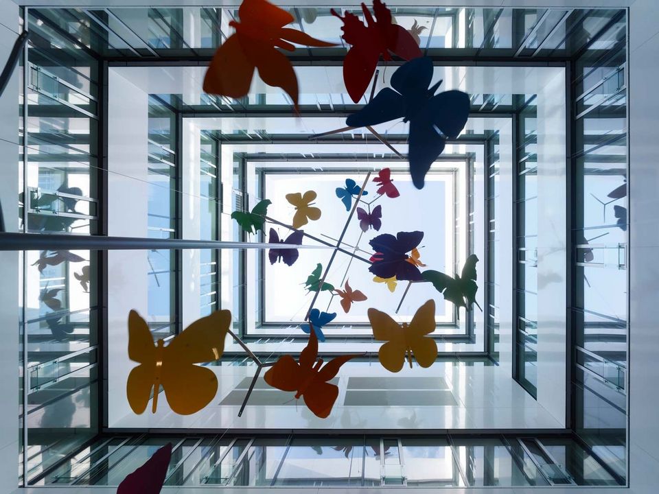 Blick nach oben durch verglastes Atrium mit hängender Installation aus bunten Schmetterlingsformen, lichtdurchflutete Architektur