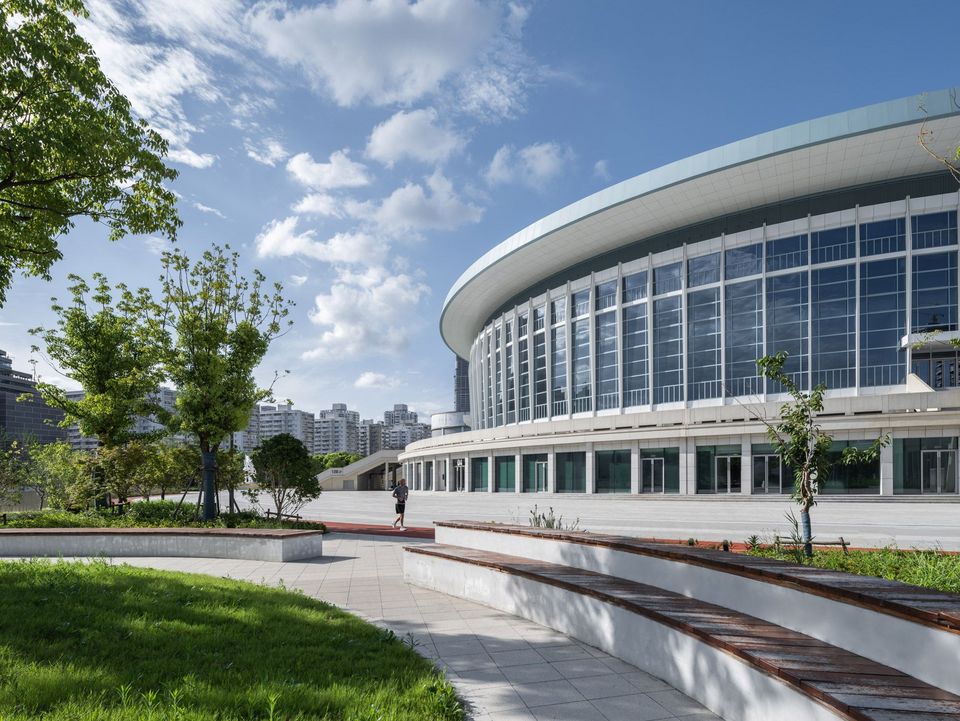 Außenaufnahme einer Arena mit rundem Baukörper, umlaufender Verglasung, breiten Gehwegen und parkähnlichen Grünflächen im Vordergrund.