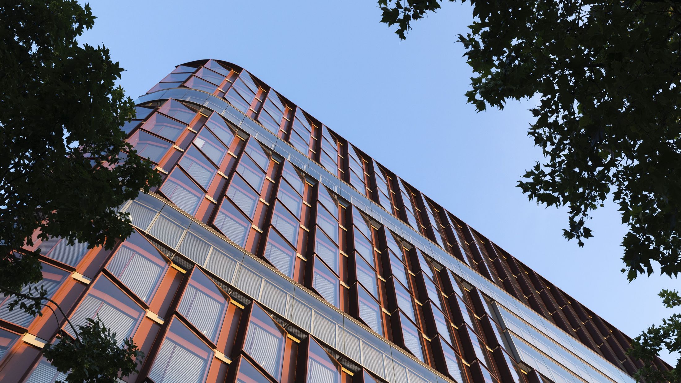 Nahaufnahme einer markanten Fassadenkante mit vertikalen, rötlich schimmernden Elementen und großformatigen Glasflächen im Seitenlicht.