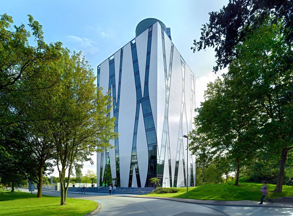 Markantes Hochhaus mit verspiegelter Glasfassade und diagonalen Streifen, umgeben von Grünflächen und Bäumen