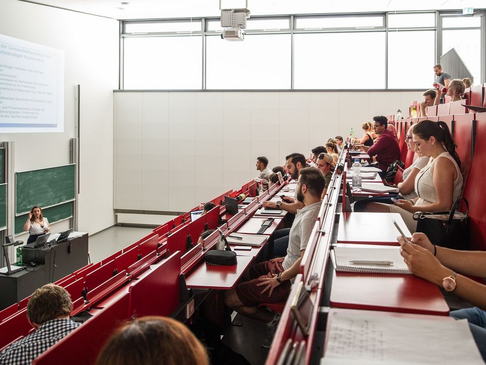 Innenaufnahme eines Hörsaals mit ansteigender Sitzreihe, Präsentationsfläche und Studierenden während der Vorlesung.