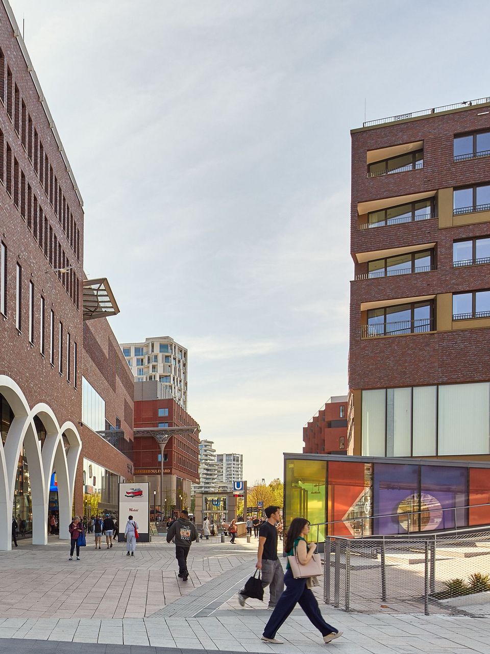 Außenaufnahme eines urbanen Straßenraums zwischen Ziegelfassaden mit Einzelhandelsbereichen, Passanten und moderner Stadtraumgestaltung.