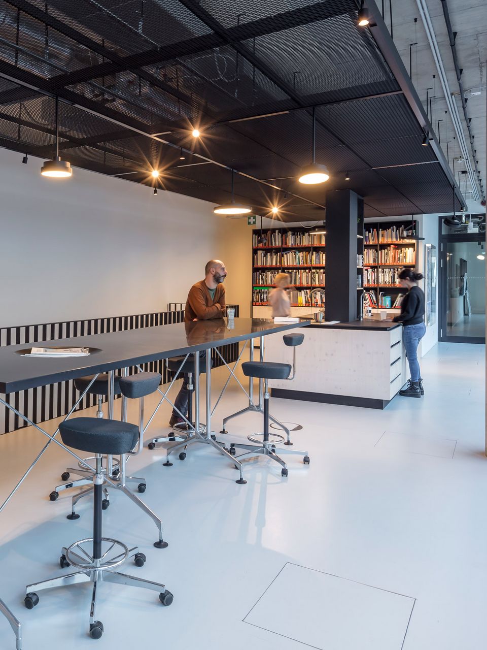 Innenaufnahme einer offenen Bibliotheks‑ und Arbeitszone mit hohen Tischen, Regalwänden und industriell gestalteter Deckenstruktur.