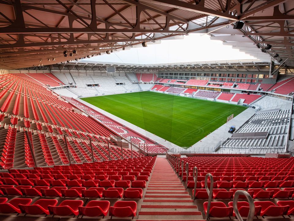Panoramablick über das Stadion mit roten Sitzreihen und grünem Spielfeld, klare Dachkonstruktion mit sichtbaren Stahlträgern