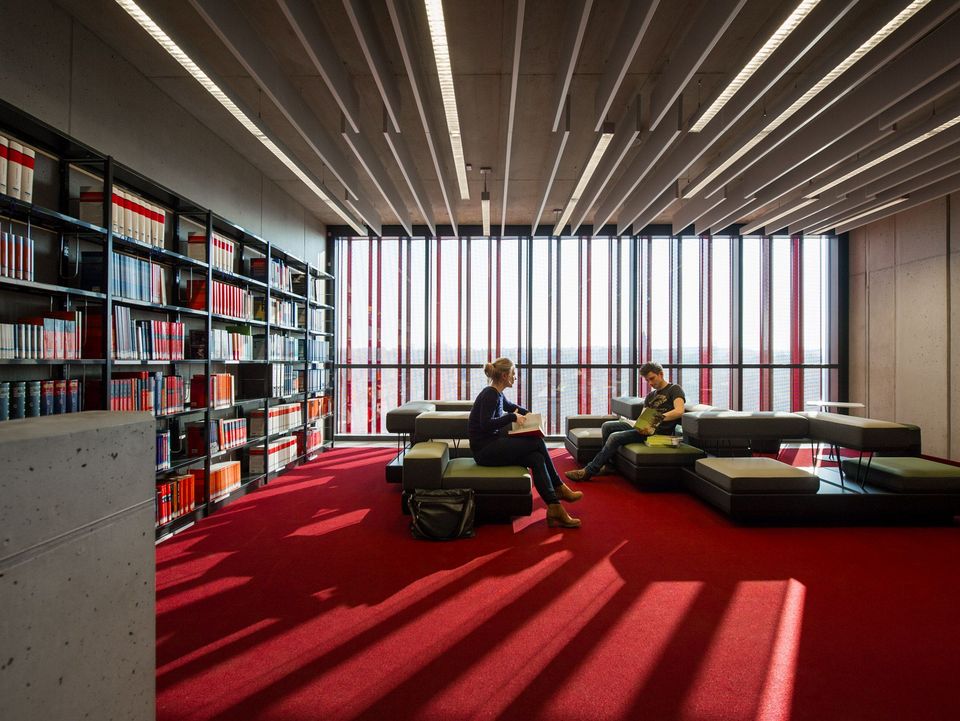 Innenaufnahme eines Bibliotheksbereichs mit Regalwänden, Sitzinseln und raumhoher Verglasung mit Lamellenstruktur.