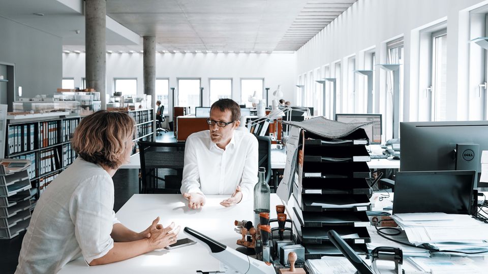 Innenaufnahme eines linearen Bürogrundrisses mit vollständig belegten Arbeitsplätzen, Monitoren, Unterlagen und Fenstern entlang der Längsseite.