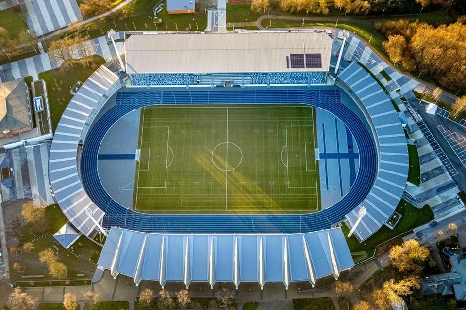 Luftaufnahme eines Stadions mit umlaufender blauer Laufbahn, zentralem Spielfeld und überdachten Tribünen.