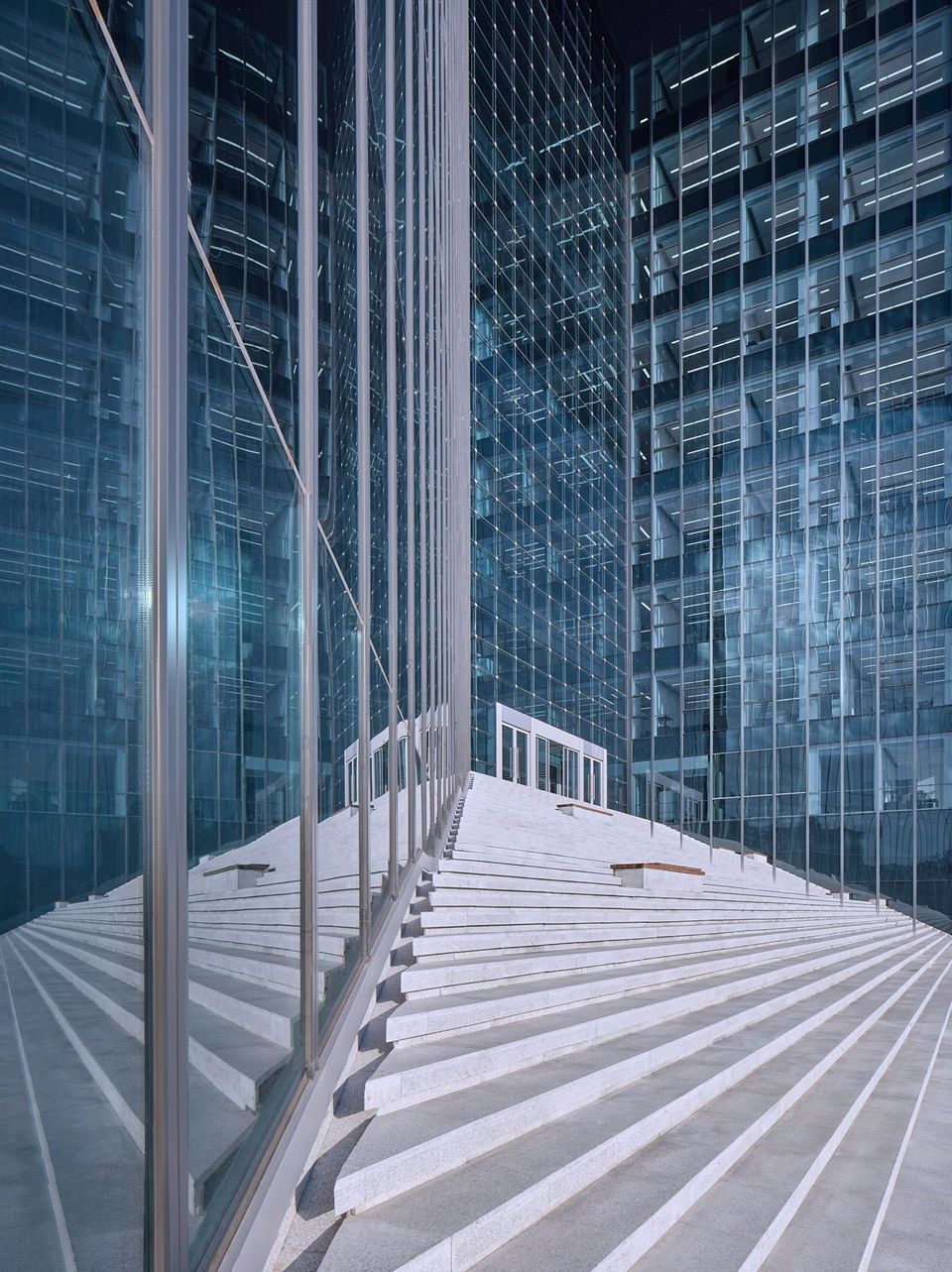Detailreiche Außenaufnahme des Eingangsbereichs mit breiter Stufenanlage, hoch aufragenden Glasfassaden und geometrischen Reflexionen, die den architektonischen Fokus auf Transparenz und Präzision betonen.
