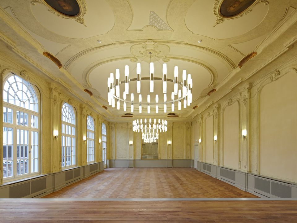 Innenaufnahme eines historischen Festsaals mit reich ornamentierter Stuckdecke, großen Rundbogenfenstern und geometrischem Parkettboden, die die denkmalgeschützte Ausstattung betonen.