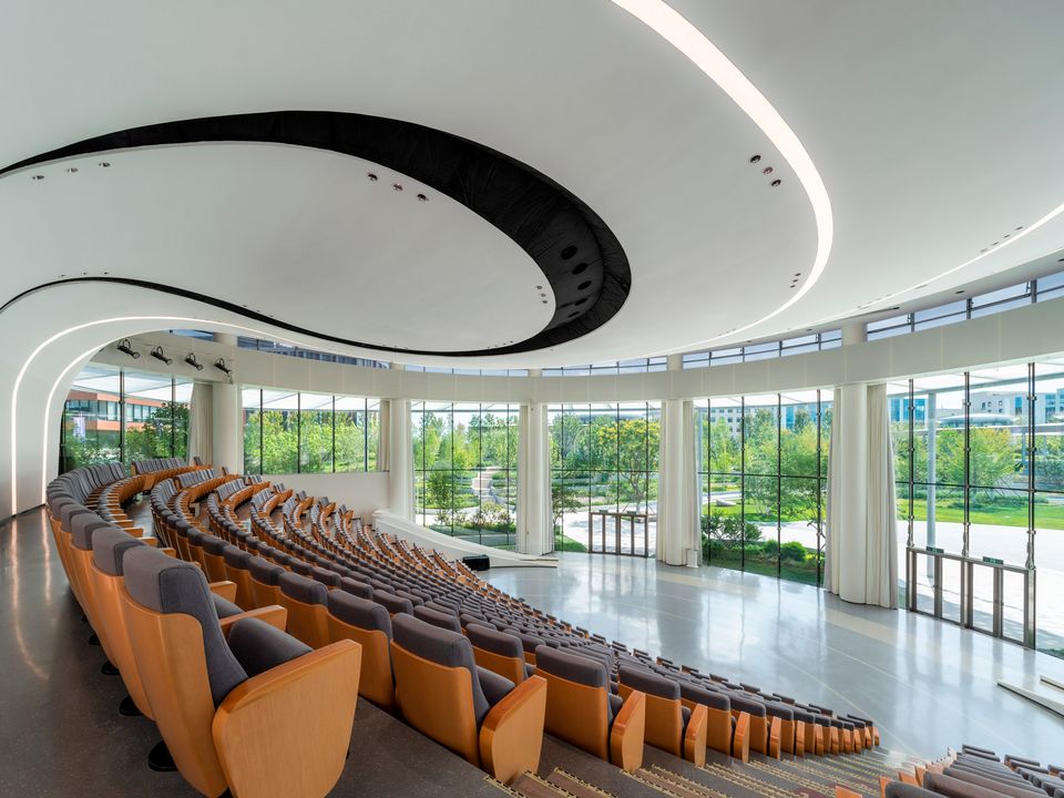 Innenaufnahme eines großzügigen Auditoriums mit geschwungener Sitzanordnung, panoramischer Glasfassade und einer dynamisch geformten Decke, die den offenen Blick in die umliegende Parklandschaft ermöglicht.