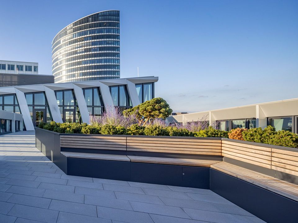 Dachterrasse mit Sitzbänken und Begrünung, Blick auf moderne Gebäudestruktur mit markanten Fassadenelementen und angrenzendem Hochhaus