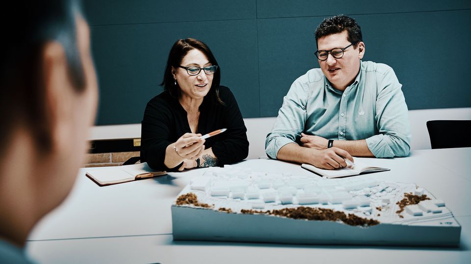 Innenaufnahme eines offenen Bürogeschosses mit mehreren Arbeitsplätzen, Regalstrukturen, Beleuchtungsschienen und architektonischen Arbeitsunterlagen.