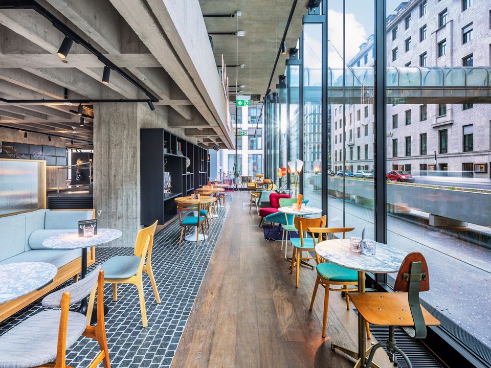 Innenaufnahme eines offenen Gastronomie‑ und Loungebereichs mit bodentiefen Glasfassaden, Sichtbetondecke, unterschiedlichen Sitzbereichen und Blick auf den umliegenden Stadtraum.