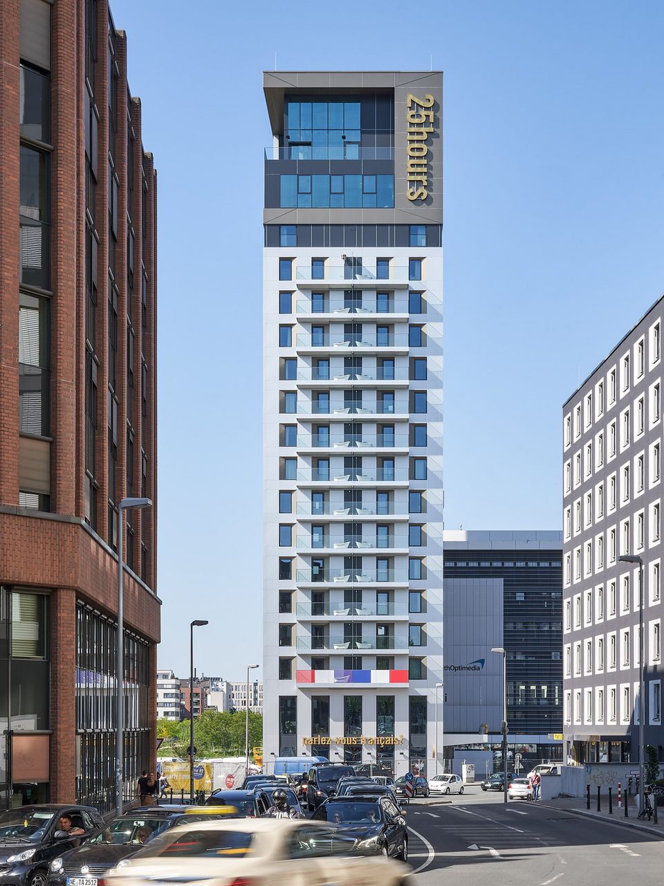 Frontalaufnahme eines schlanken Hochhauses im urbanen Straßenraum mit klar gegliederter Fassadenstruktur, auskragendem Dachabschluss und belebter Verkehrssituation im Vordergrund.