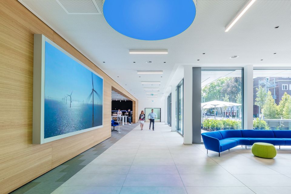 Innenaufnahme eines Foyers mit großformatigem Wanddisplay, offenem Kommunikationsbereich und verglastem Zugang zum Außenraum.