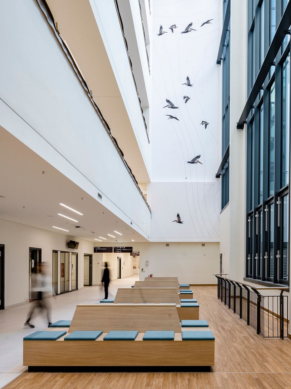 Innenaufnahme eines mehrgeschossigen Klinikfoyers mit großer Fensterfront, offenen Wartezonen und grafischer Wandgestaltung.