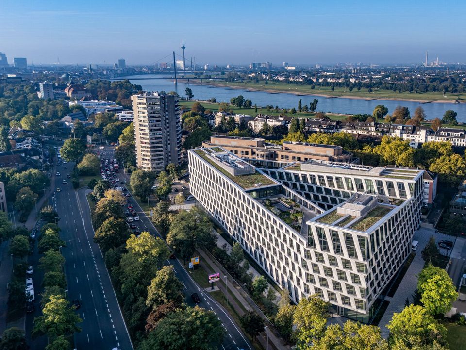 Luftaufnahme eines modernen Bürogebäudes mit markanter Fassade und begrüntem Dach mit Düsseldorfer Skyline im Hintergrund, am Rhein.
