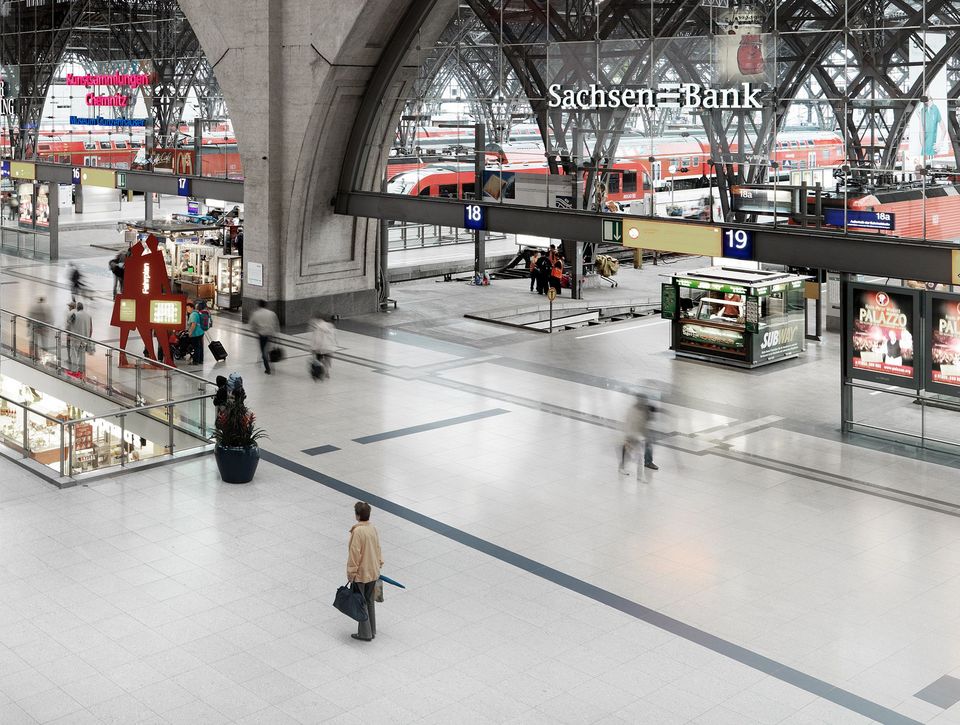 Innenraumaufnahme eines großen Kopfbahnhofs mit weit gespanntem Stahltragwerk, Bahnsteigen, Fernverkehrszügen und einem offenen Passagenbereich mit Shops und Serviceflächen im Übergang zwischen Verkehr und Handel.