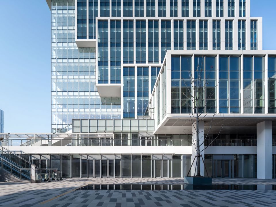Fassadendetail eines Bürogebäudes mit gläsernen Rasterstrukturen, auskragenden Ebenen und transparenter Sockelzone entlang eines weitläufigen Vorplatzes.