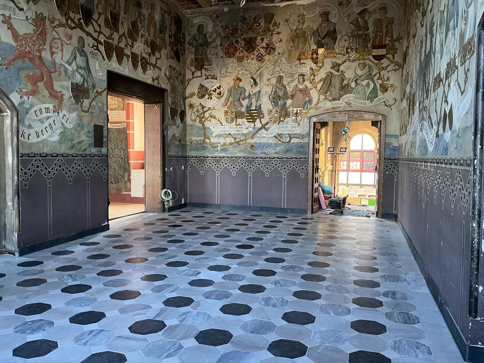 Innenaufnahme eines reich dekorierten Raums mit umlaufenden Wandmalereien, geometrischem Bodenmuster und Blick in angrenzende restaurierte Bereiche.