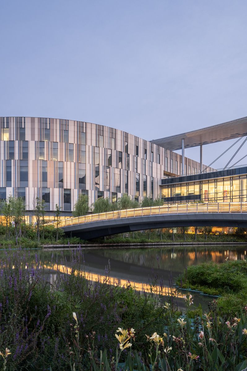 Außenaufnahme eines modernen Campusgebäudes mit vertikal strukturierter Fassade, begrüntem Uferbereich und einer verbindenden Fußgängerbrücke über ein Gewässer.