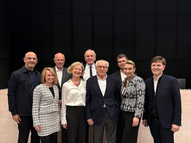 Gruppenaufnahme mehrerer Personen vor einer dunklen Wand in einem Veranstaltungsraum, aufgenommen während eines Architekturevents.