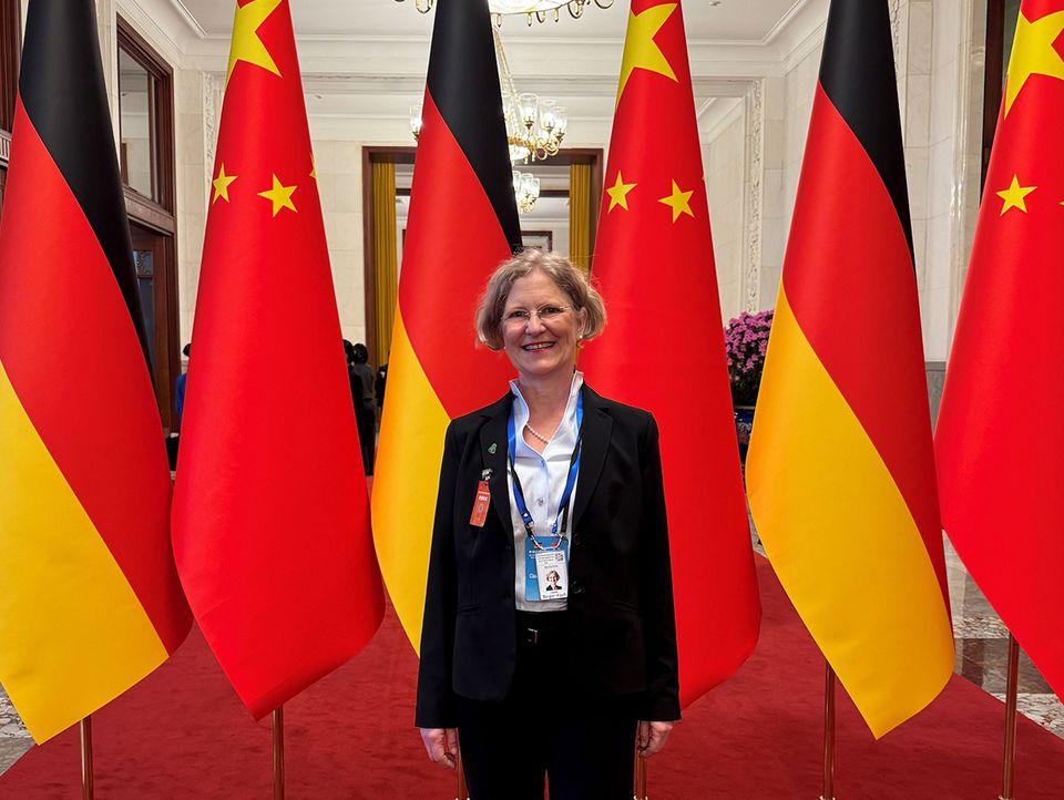 Innenaufnahme eines repräsentativen Saals mit deutschen und chinesischen Flaggen, in dem eine Person mit Delegationsausweis vor der Flaggenreihe steht.
