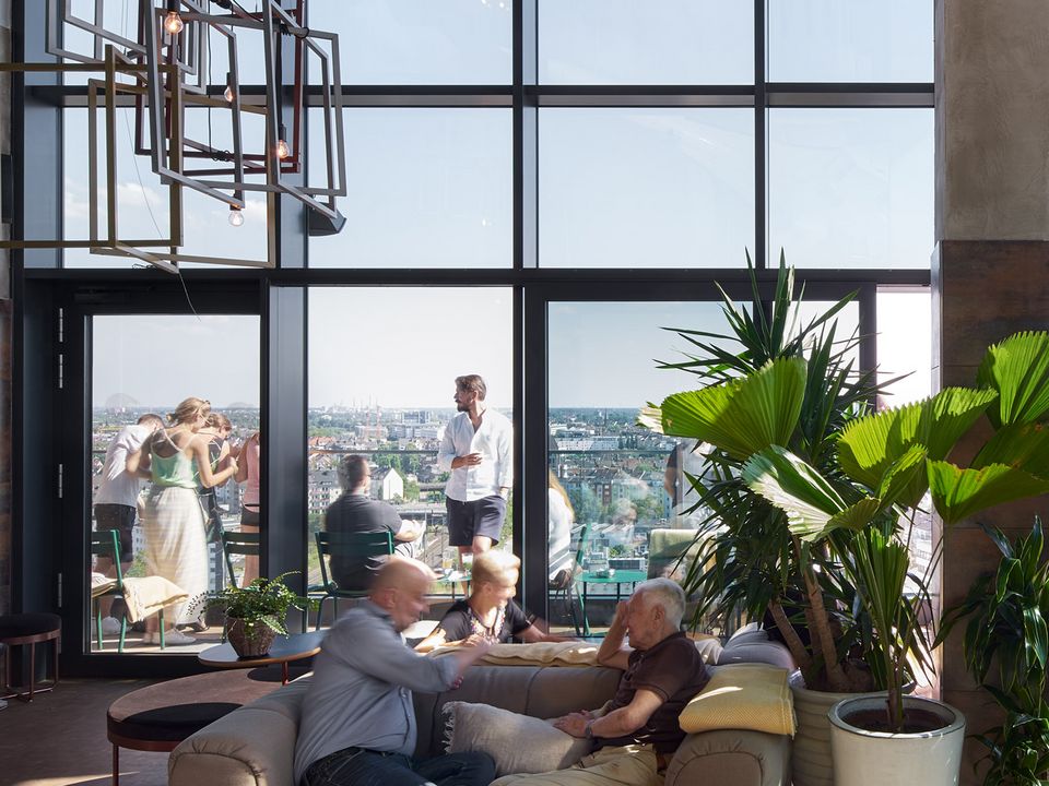 Lounge mit großen Glasfronten und Blick auf Dachterrasse, moderne Sitzmöbel und üppige Pflanzen, offene Raumgestaltung