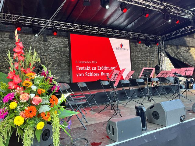 Detailaufnahme einer Bühne mit großformatiger Leinwand, floraler Dekoration und vorbereiteten Sitz- und Musikelementen für eine feierliche Veranstaltung.