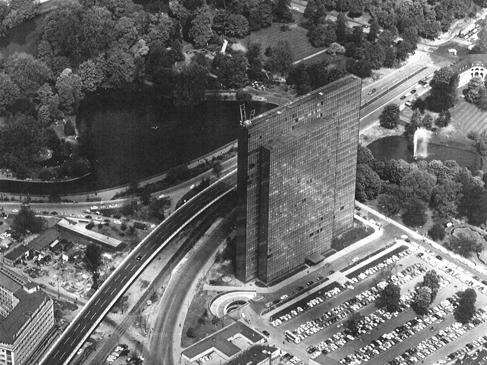 Historische Luftaufnahme eines Hochhauses mit dunkler Glasfassade, eingebettet in Grünflächen und umgeben von Straßen und Parkplätzen