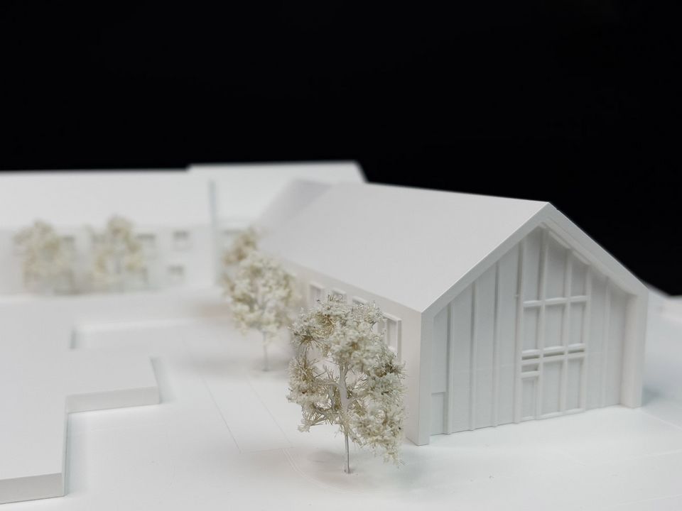 Architekturmodell eines längsorientierten Gebäuderiegels mit Giebelabschluss, umliegenden Modellbäumen und reduzierter weißer Darstellung.