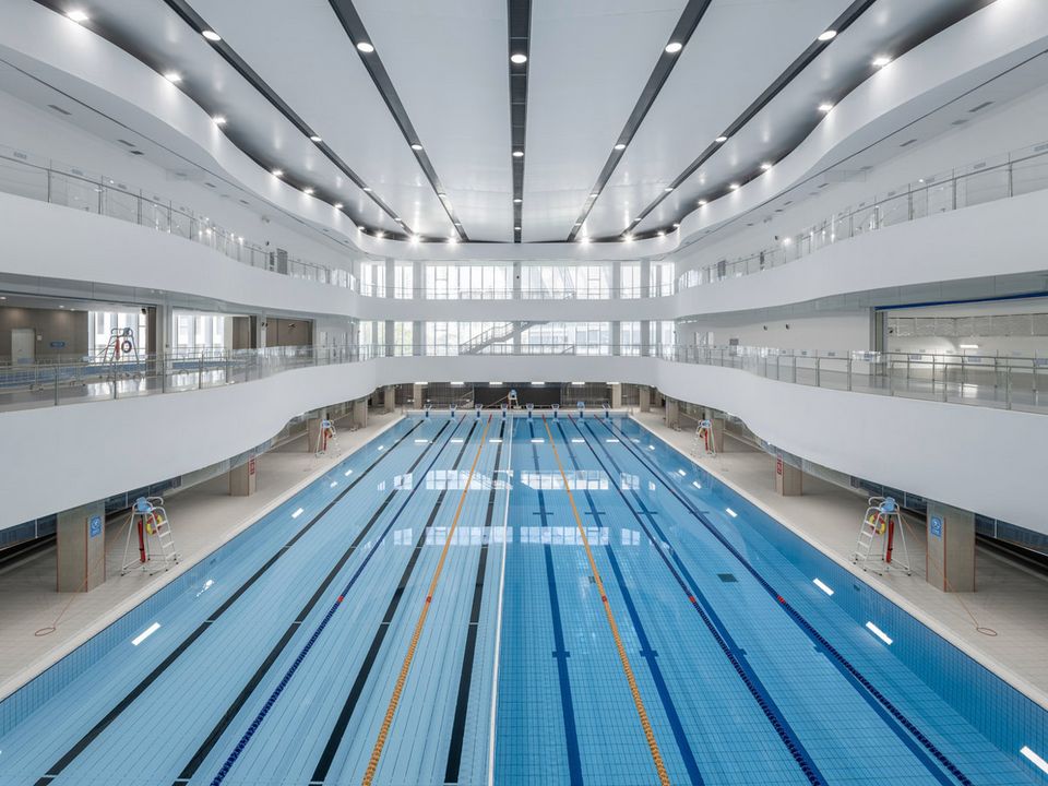 Innenaufnahme eines mehrgeschossigen Schwimmbads mit Wettkampfbecken, umlaufenden Galerieebenen, breiten Fensterfronten und klarer Linienführung.